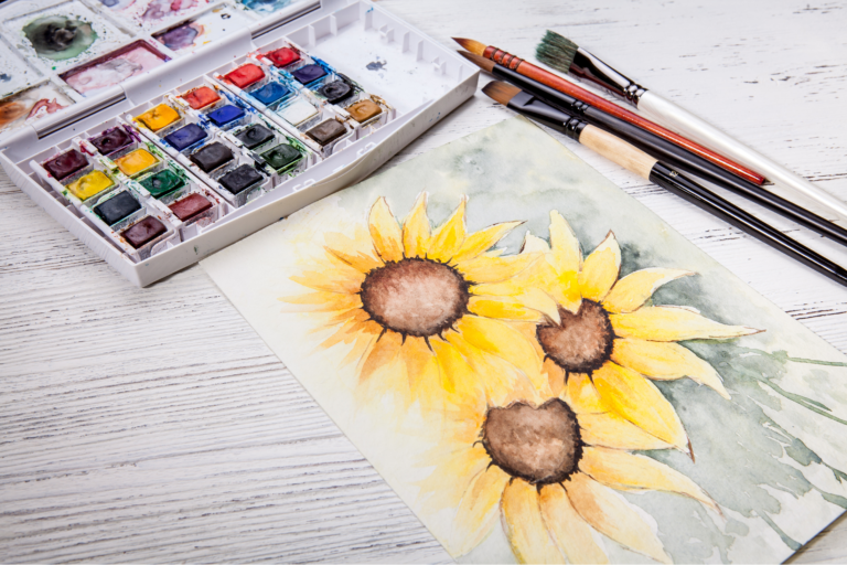 set of watercolor paints, brushes and painting of sunflowers on a table.